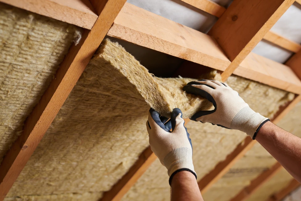 Photograph d'un artisan en tenue de travail professionnelle (salopette et t-shirt de couleur neutre) effectuant précisément un travail de rénovation intérieure tel que la pose d'enduit sur un mur, dans un environnement de chantier résidentiel. L'environnement doit montrer de l'organisation et de la propreté (protections au sol, matériaux organisés). La scène capture le sérieux et la précision de l'exécution, sans sourire artificiel ni pose forcée.
